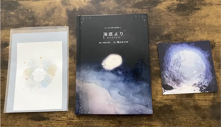 イベントで頒布したセット内容、ポストカード(左)、絵本(中)、楽曲ダウンロードカード(右)